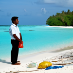 A serene panoramic view of a Maldivian island beach, featuring pristine turquoise waters and soft golden sand. Scattered along the shoreline and in the shallow water, some lightweight plastic debris, like bottles and bags, is subtly visible, indicating environmental impact without being overtly disturbing. A person, fully clothed in modest, professional attire, stands respectfully on the beach, observing the scene with a pensive yet hopeful expression. The image maintains a tranquil atmosphere, emphasizing the delicate balance between natural beauty and conservation efforts. Perfect anatomy, correct proportions, natural pose, well-formed hands, proper finger count, natural body proportions, professional photography, high quality, safe for work, appropriate content, family-friendly.